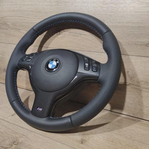 Op de afbeelding: Een zwart stuurwiel met blauwe stiksels en een geperforeerde grip. Het midden heeft een BMW-logo en koolstofvezel accenten. Het stuurwiel heeft geïntegreerde bedieningselementen en een M-logo.