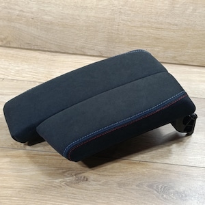 Könnte beinhalten: Ein gepolsterte, schwarze Autoarmlehne mit roten und blauen Nähten an den Kanten. Die Armlehne ist rechteckig und scheint aus einem weichen, Wildleder-ähnlichen Material zu bestehen. Der Hintergrund ist eine helle Holzoberfläche.