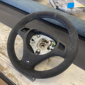 May include: A dark grey steering wheel with blue stitching and an M logo. The wheel has a suede-like texture and a multi-spoke design. The centre of the wheel reveals mechanical components. The wheel is likely for a car.