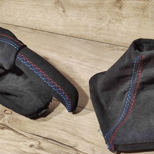 May include: Two black suede car accessories with red and blue stitching. One is a gear shift boot, and the other is a handbrake cover. The stitching is a decorative element, adding a pop of color to the dark fabric.
