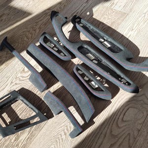May include: Various grey car interior trim components, including door handles and panels, with red and blue stitching. The parts are arranged on a wooden surface, highlighting their design and texture.