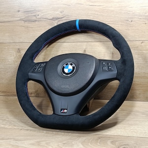 Pode incluir: Um volante preto da BMW com textura semelhante a camurça. Apresenta costuras azuis e vermelhas, uma faixa azul no topo e o logótipo da BMW no centro. A cobertura do airbag é preta e o volante tem controlos integrados.