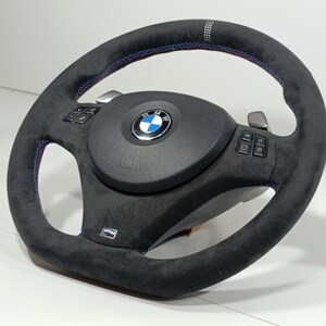 Puede incluir: Un volante negro con una textura similar a la gamuza. Cuenta con costuras azules y rojas, un logotipo de BMW y botones de control. El volante tiene un diseño deportivo.