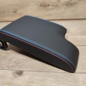 Può includere: Un componente interno automobilistico nero con cuciture rosse e blu sui bordi. L'articolo ha una forma curva e un piccolo attacco circolare su un lato. Lo sfondo è una superficie di legno marrone chiaro.