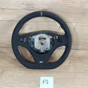 Puede incluir: Un volante negro con textura similar a la gamuza. El volante tiene un diseño de tres radios con un centro plateado y un pequeño emblema rectangular. Sobre una superficie de madera clara.