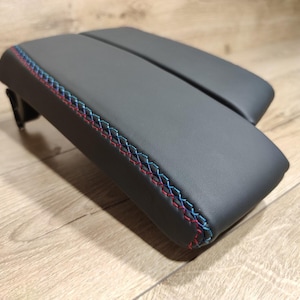 May include: A dark gray automotive armrest with red and blue stitching along the edges. The armrest is made of a smooth, leather-like material and is designed to fit in a vehicle's interior.