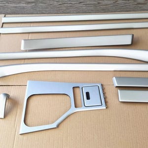 Puede incluir: Un conjunto de molduras interiores de coche de color plateado, que incluye tiras largas y rectas, piezas curvas y un panel de consola central con una abertura rectangular. Las piezas están dispuestas sobre una superficie de cartón.