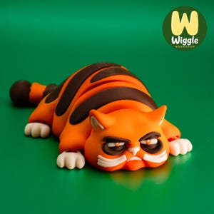 May include: An orange and brown cat figurine with a grumpy expression, white paws, and dark brown stripes. The cat is lying down on a green surface. The image includes the text "Wiggle Workshop" in a green circle.