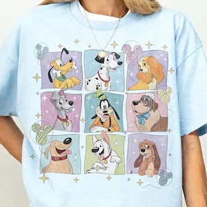 May include: Light blue t-shirt featuring a grid of cartoon dog characters. The dogs are in colorful squares with star and Mickey Mouse head accents. The shirt has short sleeves and a crew neck.