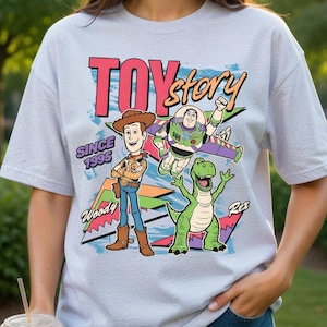 Puede incluir: Camiseta gris claro con un diseño colorido de "Toy Story", que incluye a Woody, Buzz Lightyear y Rex. La camiseta también incluye el texto "Since 1995".