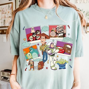 Può includere: T-shirt azzurra con una stampa colorata di personaggi dei cartoni animati. Il design include personaggi di Toy Story, Monsters & Co., Gli Incredibili e Up, con stelle e nuvole.