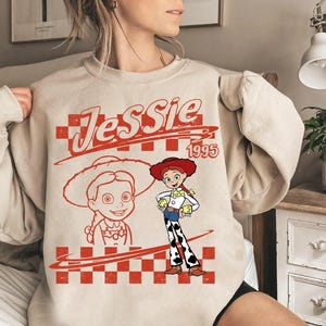 May include: A beige sweatshirt featuring a red graphic of the character Jessie from Toy Story. The design includes the name "Jessie" in a stylized font, the year "1995", and a drawing of Jessie in her cowgirl outfit.
