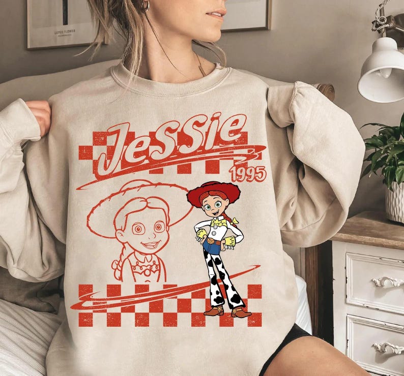 May include: A beige sweatshirt featuring a red graphic of the character Jessie from Toy Story. The design includes the name "Jessie" in a stylized font, the year "1995", and a drawing of Jessie in her cowgirl outfit.