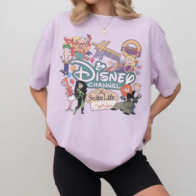 Pode incluir: Camiseta lil&aacute;s claro com um gr&aacute;fico do Disney Channel com personagens de programas como Phineas e Ferb, Hannah Montana e Kim Possible. A camiseta tem gola redonda e mangas curtas.