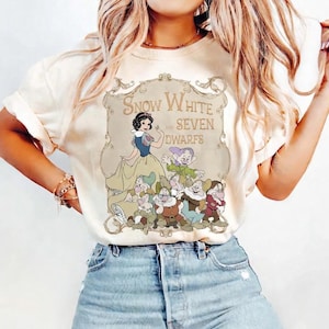 May include: Cream-colored t-shirt featuring a graphic of Snow White and the Seven Dwarfs. The design includes an illustration of Snow White in a blue and yellow dress, surrounded by the seven dwarfs, with the text "Snow White and Seven Dwarfs".