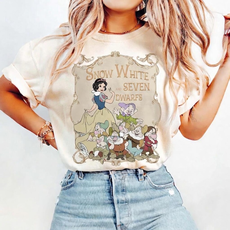 May include: Cream-colored t-shirt featuring a graphic of Snow White and the Seven Dwarfs. The design includes an illustration of Snow White in a blue and yellow dress, surrounded by the seven dwarfs, with the text "Snow White and Seven Dwarfs".