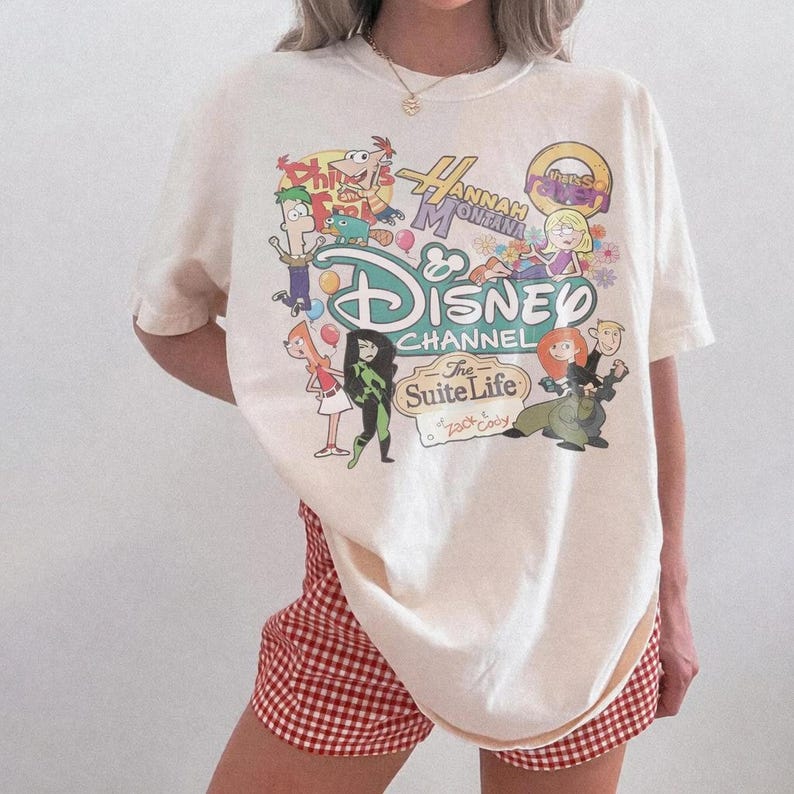 Pode incluir: Camiseta creme com um gr&aacute;fico colorido do Disney Channel com personagens de programas como Phineas e Ferb, Hannah Montana, Kim Possible e The Suite Life of Zack & Cody. Combinada com shorts xadrez vermelho e branco.