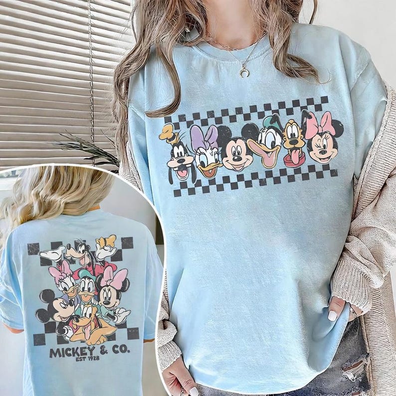 May include: Light blue t-shirt featuring a graphic of Mickey Mouse and friends, including Goofy, Donald Duck, and Minnie Mouse, set against a black and white checkered background. The back of the shirt also features a similar design with the text "Mickey & Co. EST 1928."
