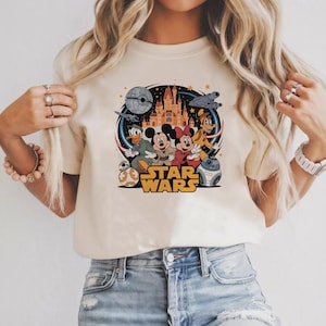Pode incluir: T-shirt creme com um gr&aacute;fico colorido que combina temas da Disney e Star Wars. O design apresenta Mickey e Minnie Mouse, Pato Donald, Pateta e outros personagens, com as palavras "STAR WARS" em dourado.