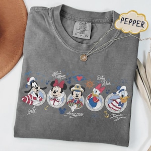 May include: A gray Comfort Colors t-shirt featuring Disney characters Goofy, Minnie Mouse, Mickey Mouse, Daisy Duck, and Donald Duck in nautical-themed designs. The shirt has a crew neck and a small tag that says "S - P - CH". A silver necklace with a pendant is also visible.