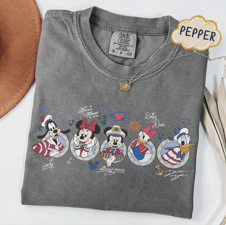 May include: A gray Comfort Colors t-shirt featuring Disney characters Goofy, Minnie Mouse, Mickey Mouse, Daisy Duck, and Donald Duck in nautical-themed designs. The shirt has a crew neck and a small tag that says "S - P - CH". A silver necklace with a pendant is also visible.