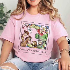 May include: A pale pink t-shirt with a framed graphic of Toy Story characters, including Woody, Buzz Lightyear, and Rex. The text "YOU'VE GOT A FRIEND IN ME" is below the graphic. The shirt is made of a soft material.