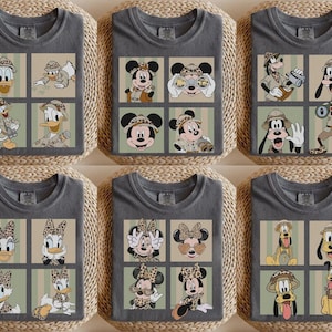 May include: Gray t-shirts featuring cartoon characters in safari attire. The shirts showcase Donald Duck, Mickey Mouse, Goofy, and Daisy Duck in various poses within a grid design. The characters are wearing safari hats and holding binoculars.