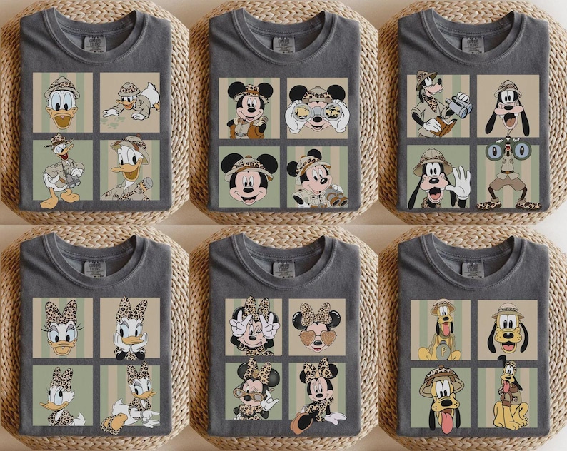 May include: Gray t-shirts featuring cartoon characters in safari attire. The shirts showcase Donald Duck, Mickey Mouse, Goofy, and Daisy Duck in various poses within a grid design. The characters are wearing safari hats and holding binoculars.