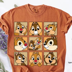 Op de afbeelding: Verbrand oranje t-shirt met een raster van negen vierkanten, elk met een ander cartoon eekhoorn gezicht. De eekhoorns hebben bruine en bruine vacht, rode neuzen en verschillende uitdrukkingen. Het shirt is gemaakt van een zacht materiaal.