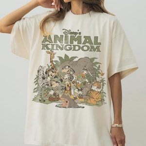 K&ouml;nnte beinhalten: Cremefarbenes T-Shirt mit dem gr&uuml;nen Schriftzug "Disney's Animal Kingdom" &uuml;ber einer Grafik von Disney-Figuren als Safaritiere. Die Grafik zeigt ein Zebra, eine Giraffe, Micky Maus und Donald Duck.