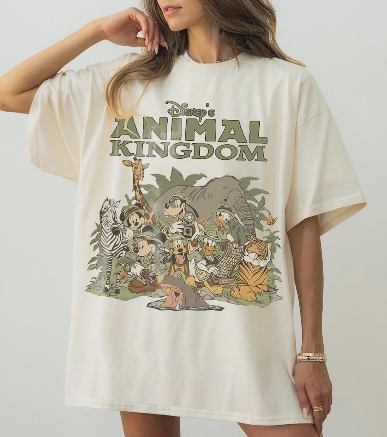 K&ouml;nnte beinhalten: Cremefarbenes T-Shirt mit dem gr&uuml;nen Schriftzug "Disney's Animal Kingdom" &uuml;ber einer Grafik von Disney-Figuren als Safaritiere. Die Grafik zeigt ein Zebra, eine Giraffe, Micky Maus und Donald Duck.