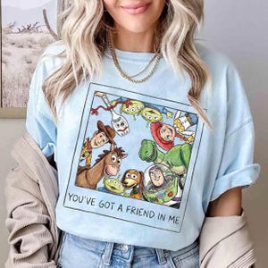 May include: Pale blue t-shirt with a colourful graphic of Toy Story characters. The design includes Woody, Buzz Lightyear, and others, with the words "YOU'VE GOT A FRIEND IN ME" printed below the image.