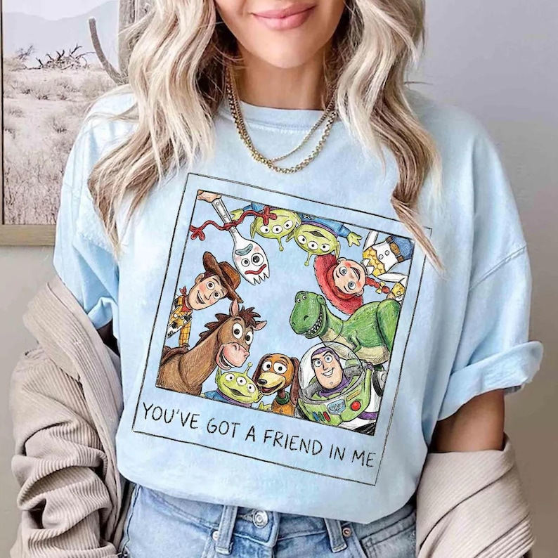 May include: Pale blue t-shirt with a colourful graphic of Toy Story characters. The design includes Woody, Buzz Lightyear, and others, with the words "YOU'VE GOT A FRIEND IN ME" printed below the image.