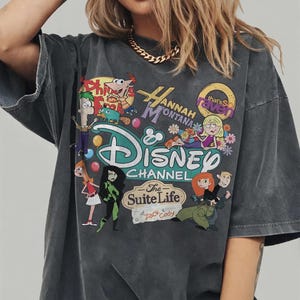 Pode incluir: T-shirt cinza escuro com um design gr&aacute;fico colorido com as palavras "Hannah Montana" e "Disney Channel", juntamente com personagens de desenhos animados. A camiseta tem mangas curtas e um corte folgado.