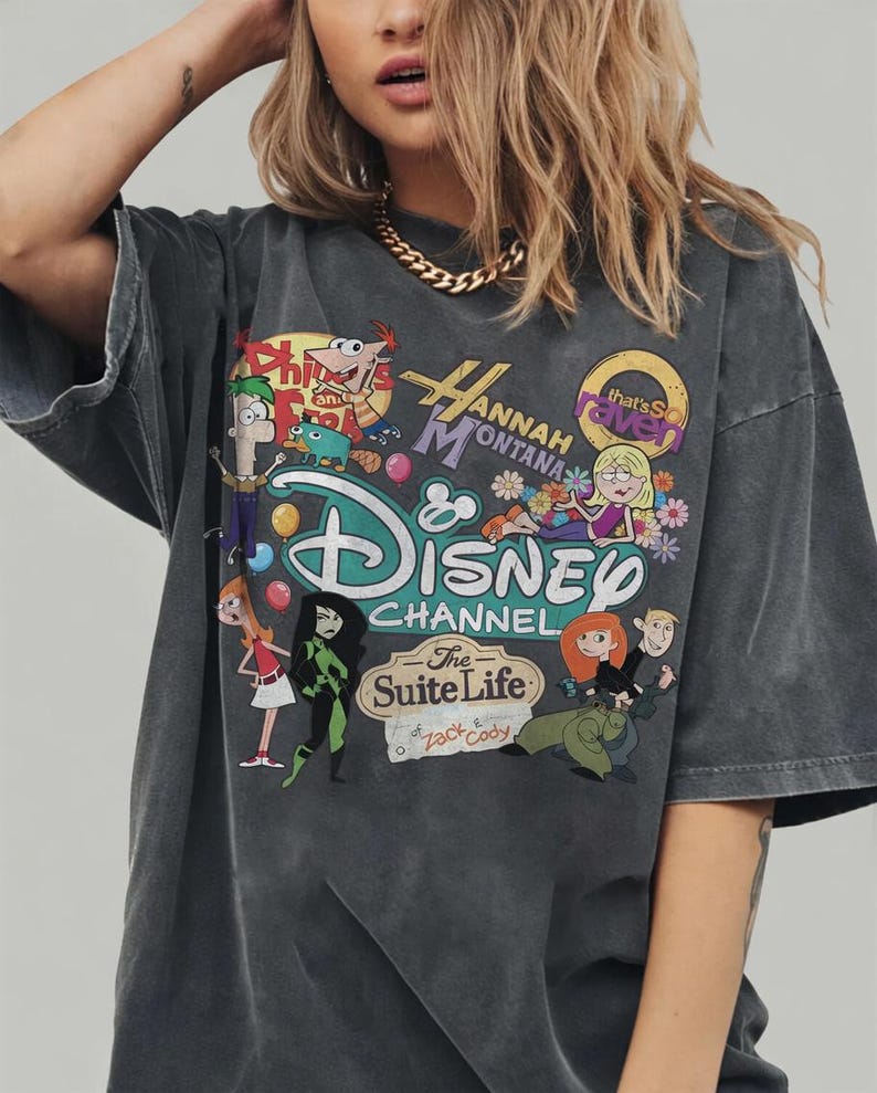 Pode incluir: T-shirt cinza escuro com um design gr&aacute;fico colorido com as palavras "Hannah Montana" e "Disney Channel", juntamente com personagens de desenhos animados. A camiseta tem mangas curtas e um corte folgado.