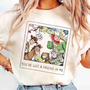 Könnte beinhalten: Cremefarbenes T-Shirt mit einer farbenfrohen Illustration von Charakteren aus dem Animationsfilm "Toy Story". Das Design zeigt Woody, Buzz Lightyear, Rex und andere Charaktere in einem Polaroid-Rahmen mit dem Text "YOU'VE GOT A FRIEND IN ME."