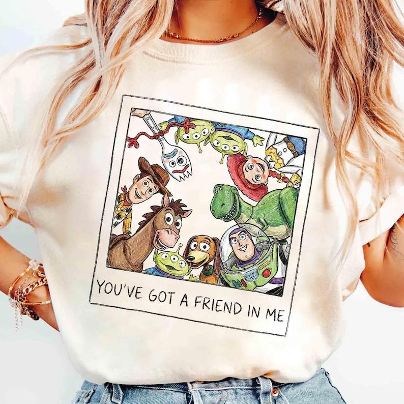 May include: A cream-coloured t-shirt with a colourful illustration of characters from the animated film "Toy Story." The design includes Woody, Buzz Lightyear, Rex, and other characters within a polaroid frame, with the text "YOU'VE GOT A FRIEND IN ME."