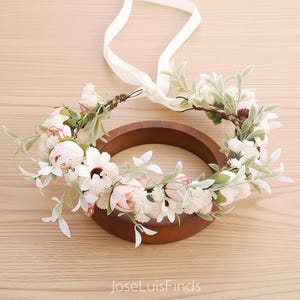 Puede incluir: Una corona floral con flores blancas y rosas, hojas verdes y una cinta blanca. La corona se exhibe sobre un anillo de madera. El texto "JoseLuisFinds" es visible en la parte inferior.