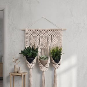 May include: A cream-coloured macrame wall hanging featuring three integrated plant holders suspended from a wooden dowel. The piece displays intricate knotted patterns and includes three tassels at the bottom, each holding a potted plant.