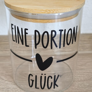 Könnte beinhalten: Ein klares Glas mit einem Bambusdeckel. Das Glas hat schwarzen Text, der "EINE PORTION GLÜCK" mit einem Herzsymbol liest. Das Glas steht auf einer Holzoberfläche.