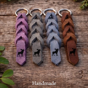 Op de afbeelding: Vier handgemaakte Nordic lederen sleutelhangers in paars, grijs, blauwgrijs en bruin. Elke sleutelhanger heeft een gevlochten ontwerp en een zwart eland silhouet. De sleutelhangers hebben zilveren metalen ringen.