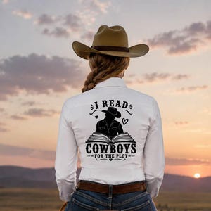 May include: A white long-sleeve shirt featuring a black graphic on the back with a cowboy silhouette, an open book, and the text I READ COWBOYS FOR THE PLOT. The shirt is worn with blue denim jeans and a brown cowboy hat while riding a brown horse.