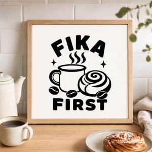 Könnte beinhalten: Ein quadratischer Holzrahmen mit einem weißen Schild, auf dem in schwarzer Schrift FIKA FIRST steht. Das Design zeigt eine dampfende Kaffeetasse, eine Zimtschnecke und Kaffeebohnen. Die Grafik ist mittig auf weißem Grund im Rahmen platziert.