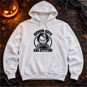 Könnte beinhalten: Ein weißer Kapuzenpullover mit einem schwarzen Grafikdruck einer Puppe mit zerzaustem Haar und gestreifter Kleidung in einem Kreis. Der Text GOOD GUY ist darüber gebogen und BAD ATTITUDE ist in fetten schwarzen Buchstaben darunter gedruckt.