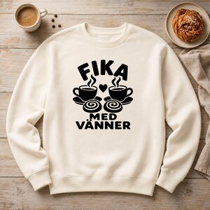 Könnte beinhalten: Ein cremefarbenes Sweatshirt mit Rundhalsausschnitt und einem schwarzen Grafikdruck auf der Brust. Das Design zeigt zwei dampfende Kaffeetassen, zwei Zimtschnecken, ein kleines Herz und den Text FIKA MED VÄNNER in einer fetten Schriftart.