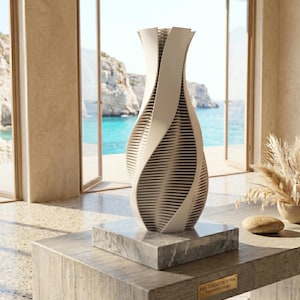 Könnte beinhalten: Eine weiße und beige Spirale Vase mit einem einzigartigen Design, auf einem Marmorsockel. Die Vase steht auf einem Betontisch mit einem Küstenhintergrund aus Meer und Klippen. Die Vase trägt den Text "AVS TURQUOISE SPIRAL Parametric Design".