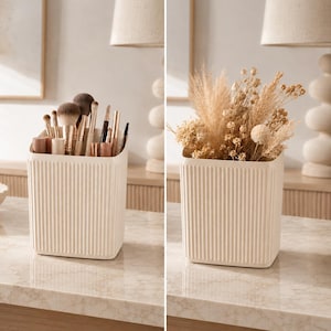 May include: A cream-colored, ribbed container, shown with makeup brushes and dried flowers. The container has a rectangular shape with vertical lines. The brushes are various sizes and colors. The dried flowers are beige and tan.