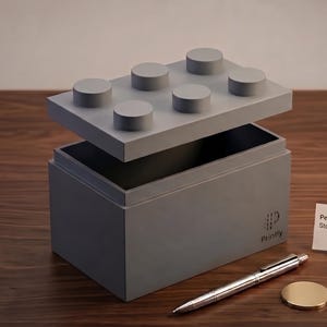 Può includere: Un cubo contenitore grigio con un coperchio progettato per assomigliare a un grande mattoncino. Una penna d'argento e una moneta d'oro sono accanto al cubo. Una piccola carta recita "Personalized Large-Scale Storage Cube STL File."