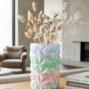 May include: A pastel-colored geometric vase holds dried flowers. The vase transitions from blue to green to pink. The flowers are beige and white, adding a natural element to the modern decor. A gold pen rests on a white tray.