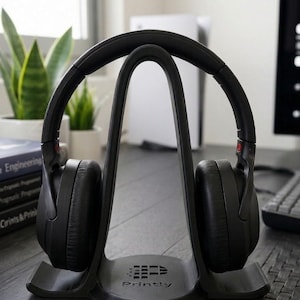 May include: Black over-ear headphones resting on a dark gray headphone stand. The stand has a curved design with the word "Printly" printed on the base. The headphones have red accents and are positioned on a wooden surface.